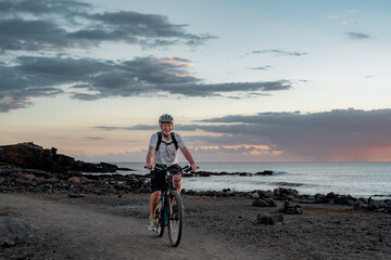 Happy active senior cyclist man at sea at sunset light with electric bicycle running on the beach -...