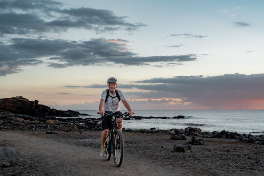 Happy active senior cyclist man at sea at sunset light with electric bicycle running on the beach - elderly man with helmet enjoying healthy lifestyle and freedom