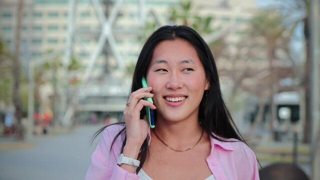 Slow motion fotage of one asian woman walking and having a friendly smart phone call conversation. Chinese pretty female teenage student smiling and talking by cellphone. Girl speaking by mobile phone