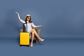Happy Asian woman traveler sitting on armchair with yellow suitcase isolated on blue background,...