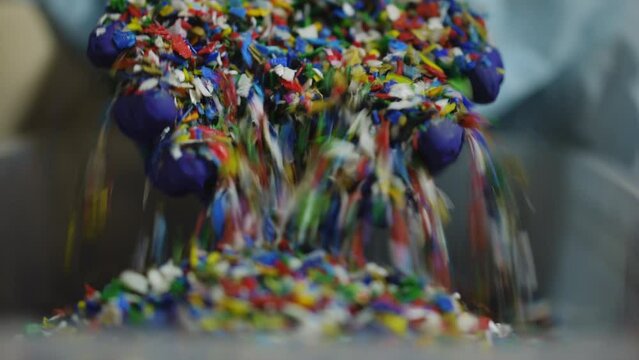 Close-up shredded multicolored plastic garbage with female hands in gloves raising smashed trash showing colorful parts falling in box. Recycling and waste separation concept