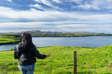Back shot of a young brown-haired Caucasian woman by a fence calmly observing the beautiful landscape in the Scottish Highlands, with a loch in the distance. Copy space.