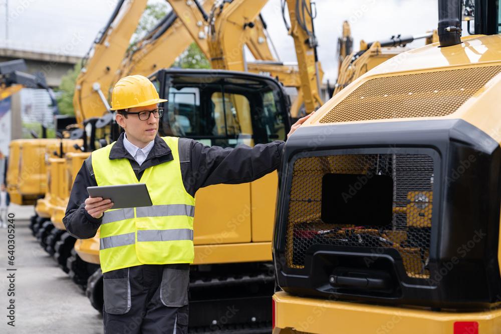 Poster engineer in a helmet with a digital tablet stands next to construction excavators.