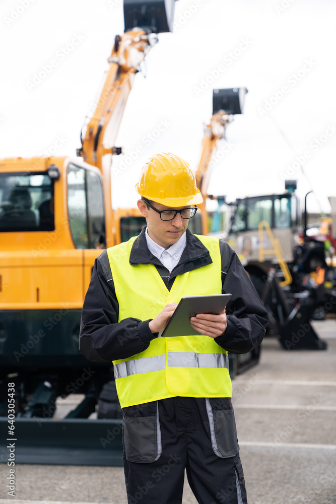 Poster engineer in a helmet with a digital tablet stands next to construction excavators.