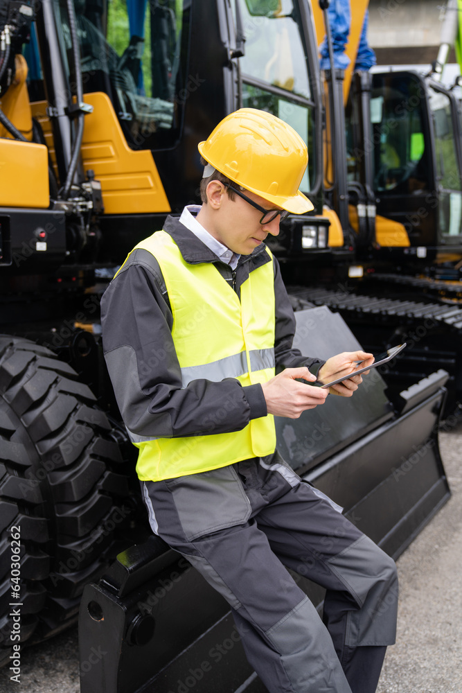 Poster engineer in a helmet with a digital tablet stands next to construction excavators.