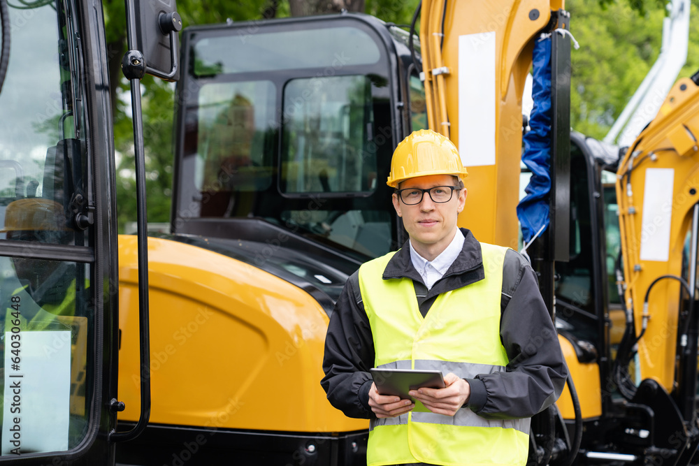 Poster engineer in a helmet with a digital tablet stands next to construction excavators.