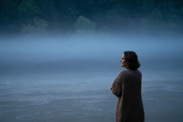 A woman in a sweater on the bank of a foggy river in the evening