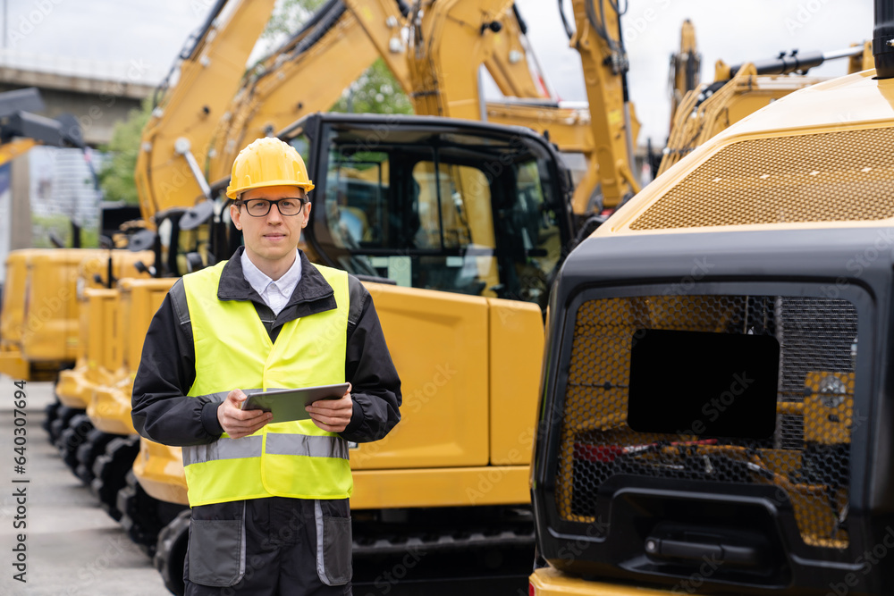 Poster engineer in a helmet with a digital tablet stands next to construction excavators.