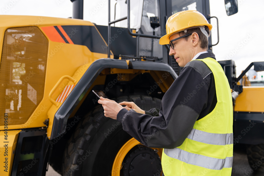 Poster Worker in a helmet with a digital tablet on the background of construction machine.