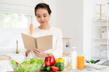 Obraz na płótnie Canvas Smiling housewife woman looking at recipe in cookery book preparing vegetable salad cooking food in light kitchen at home. Dieting healthy lifestyle concept.