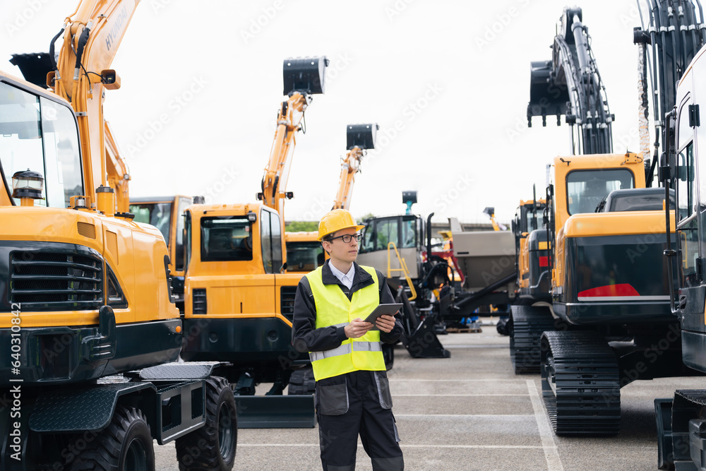 Poster engineer in a helmet with a digital tablet stands next to construction excavators.