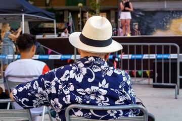 Older man with a hat and flower shirt, sitting in the park at a music concert.