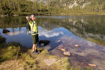 Fototapeta na wymiar A man with a backpack stands with his hands raised. against the backdrop of a mountain lake with a beautiful reflection