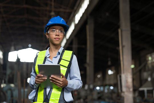 Portrait African American female engineer with tablet working in heavy metal industrial factory. Confident technician woman wearing vest and helmet safety standing at the manufacturing plant.