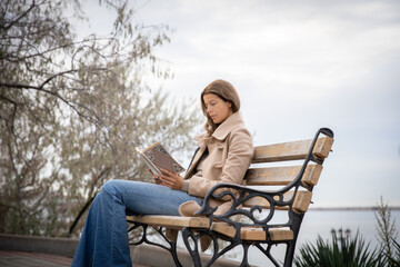 Fototapeta premium Young beautiful slim woman in a light coat and blue jeans is reading a book on a bench in the city.