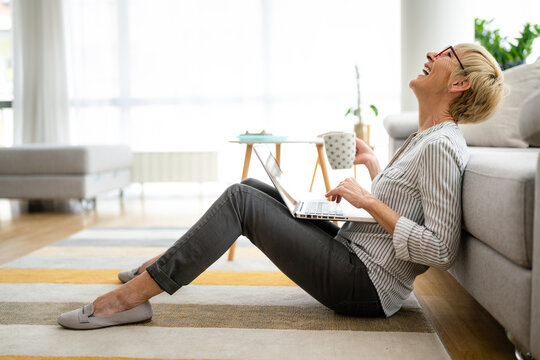 Thrilled excited laughing elderly short hair woman drinking coffee while chatting online using laptop.