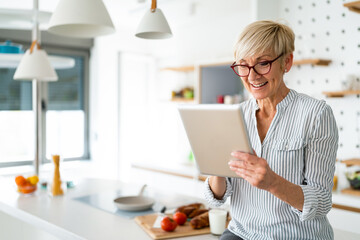Portrait of beautiful happy older woman looking for homemade recipes on digital tablet device while sitting in kitchen.