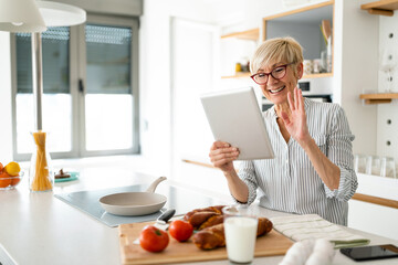 Content jovial elderly short hair woman saying hello to her friends on digital tablet while cooking healthy meal in her kitchen.