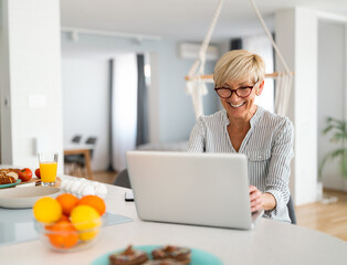 Smiling cheerful blonde hair woman with glasses, texting online via laptop, checking e-mails, using laptop for online banking.