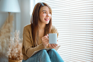 Joyful young asian female enjoying a cup of coffee while sitting on bench at home, Cosy scene,...