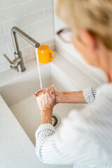 Photo from behind of unrecognizable elderly blonde woman washing hands with soap at sink in kitchen.