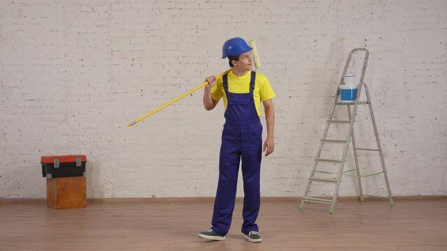 Full length video of a smiling young worker standing in the room, placing the roller at his shoulder, looking at freshly painted walls and giving a thumbs up.