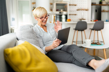 Joyous blonde senior woman waving at digital tablet while speaking to her family and sitting with...