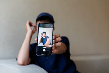teenage boy holding a smart phone in front of his face at home, taking selfies with his smartphone, digital display on foreground, teen with his own portrait in smartphone, teen blogging - obrazy, fototapety, plakaty