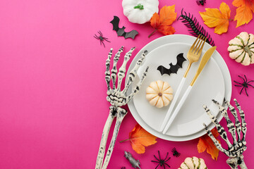 Beautiful table arrangement for Halloween. Top view composition of plates, cutlery, pumpkins, spooky skeleton hands, autumn leaves, creepy decorations on vivid pink background with promotional slot