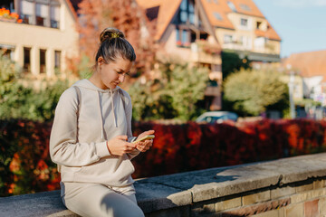 Obraz premium Young Fashionable Teenage Girl With Smartphone In Europian Park In Autumn Sitting At Smiling. Trendy Young Woman In Fall In Park Texting. Retouched, Vibrant Colors. Beautiful Blonde Teenage Girl