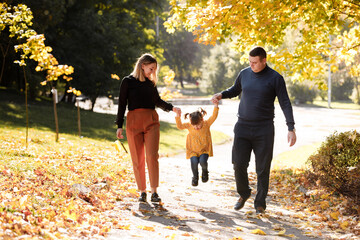 Family day. Happy smiling dad and mom having fun with their child daughter outdoors at autumn park. Couple lifting their cheerful girl kid up by the hands, enjoying nature and walking. Children's day