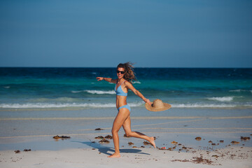 Portrait of slim woman running on the beach in a bikini and a straw hat in hand.