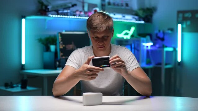 Portrait of focused young man looking, examining, testing out professional new mini LED lamp sitting at desk on background of desktop computer with neon light background. Shooting in slow motion.
