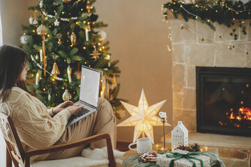 Woman in cozy sweater working on laptop with empty screen on background of christmas tree lights in stylish festive decorated room. Christmas shopping online and remote work