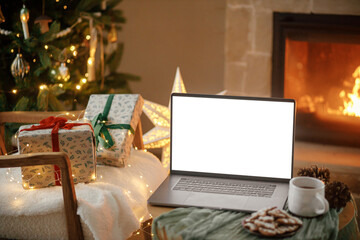 Christmas shopping online and sales. Laptop with empty screen and stylish christmas gifts on background of fireplace, tree with golden lights in festive cozy room. Laptop mock up, copy space