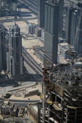 Cranes loom over the top of an building that is under construction, rising up over the skyline of Dubai. The aerial view gives a dizzying sense of the height of the new skyscrapers.