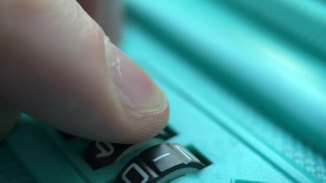 Man's finger dials the code on the lock of a turquoise polycarbonate travel suitcase. Extreme close-up