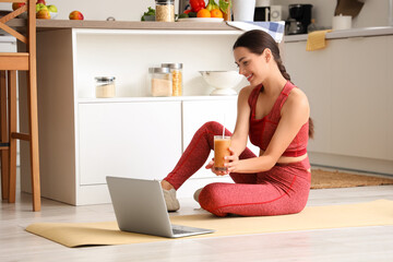 Sporty young woman with healthy smoothie and laptop in kitchen