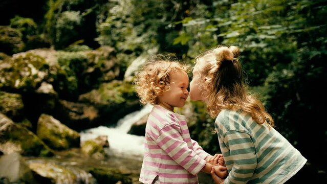 Little sisters kiss and hug each other at beautiful Alpine waterfall in the woods