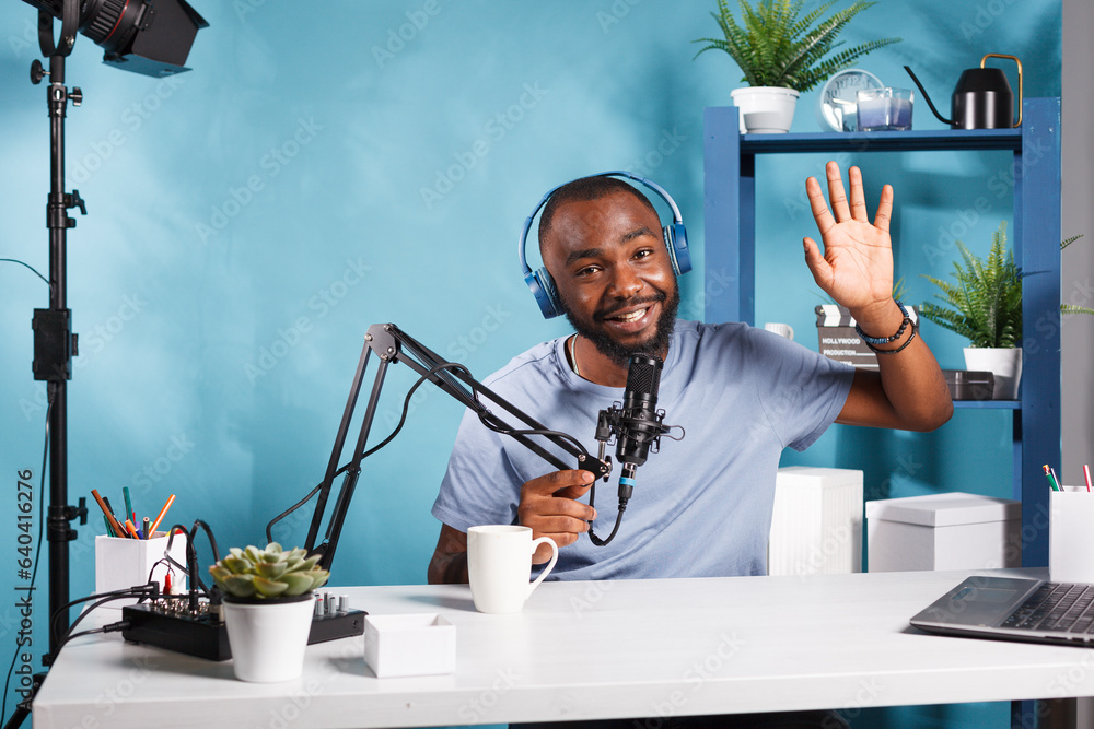 Poster smiling streamer wearing headphones greeting audience while creating digital content and looking at 