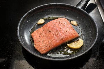 Fresh salmon filet is fried on the skin with lemon, herbs and garlic in a black frying pan on the stovetop, copy space, selected focus