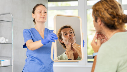 Adult man carefully scrutinizing his own reflection in mirror held by professional female cosmetologist or plastic surgeon, contemplating potential improvements or changes to appearance..