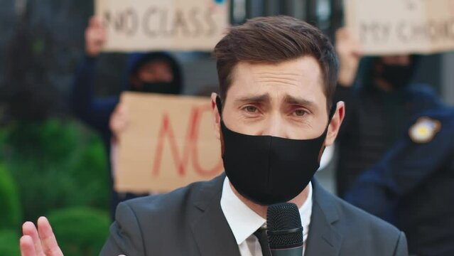 Close up portrait of young journalist in protective mask broadcasts live from scene. Report for live television from demonstration. Сrowd of people protest against vaccination and coronavirus.