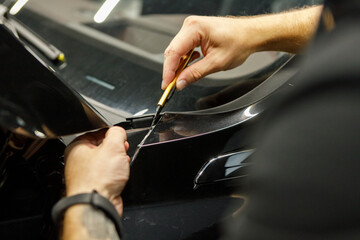 Detailer worker cuts with knife wrapped and installed PPF for car protection film on vehicle. Transparent film. Car paint protection - obrazy, fototapety, plakaty