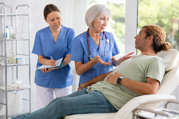 Obraz premium Female nurse and mature woman doctor carries out questionnaire survey of male patient about allergic reactions and intolerance to drugs for anesthesia