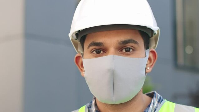 Close up portrait of young confident arhitect in protective mask with blueprint. Professional worker in yellow vest and helmet standing outdoors and looking at camera. Safety constructing concept.