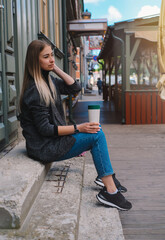 A beautiful caucasian girl with blond hair in a black jacket and blue jeans works with a thermo mug with coffee or tea on the street in clear weather.