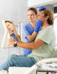 Positive adult man looking at mirror while skilled female cosmetologist showing results of hardware...