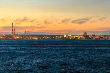 City lights and smokestack viewed from offshore with sunset glow in sky