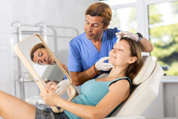 Obraz premium Interested adult female patient sitting on couch in medical aesthetic office looking at mirror while professional male cosmetologist explaining future hardware rejuvenating facial procedure